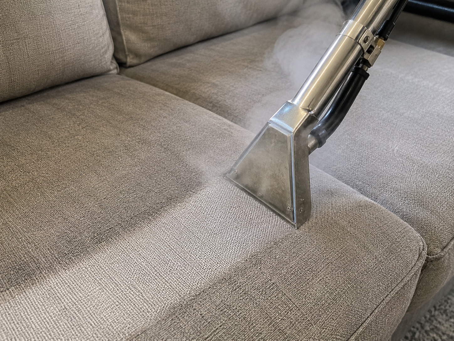 Hot-water extraction wand cleaning a fabric couch cushion with visible steam, illustrating the deep-cleaning process